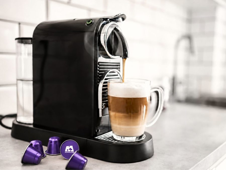 Une machine à capsules noire dans une cuisine, un café qui coule dans un verre, des capsules Mush Caps à côté